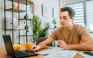 A young man working on a laptop at a desk with papers, analyzing real estate market data while holding a pen, with a modern home interior in the background.